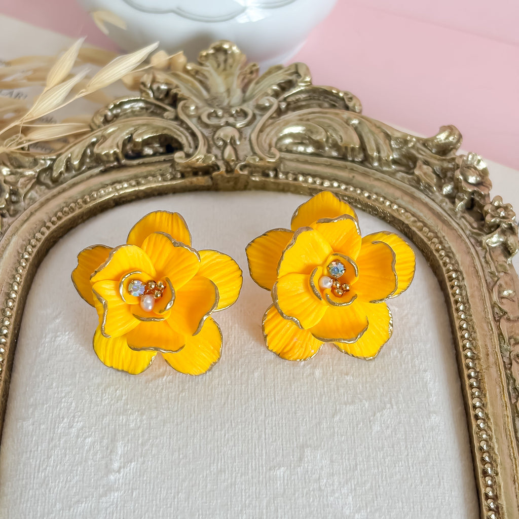 Yellow flower earrings on a white surface with a decorative gold frame and pink background