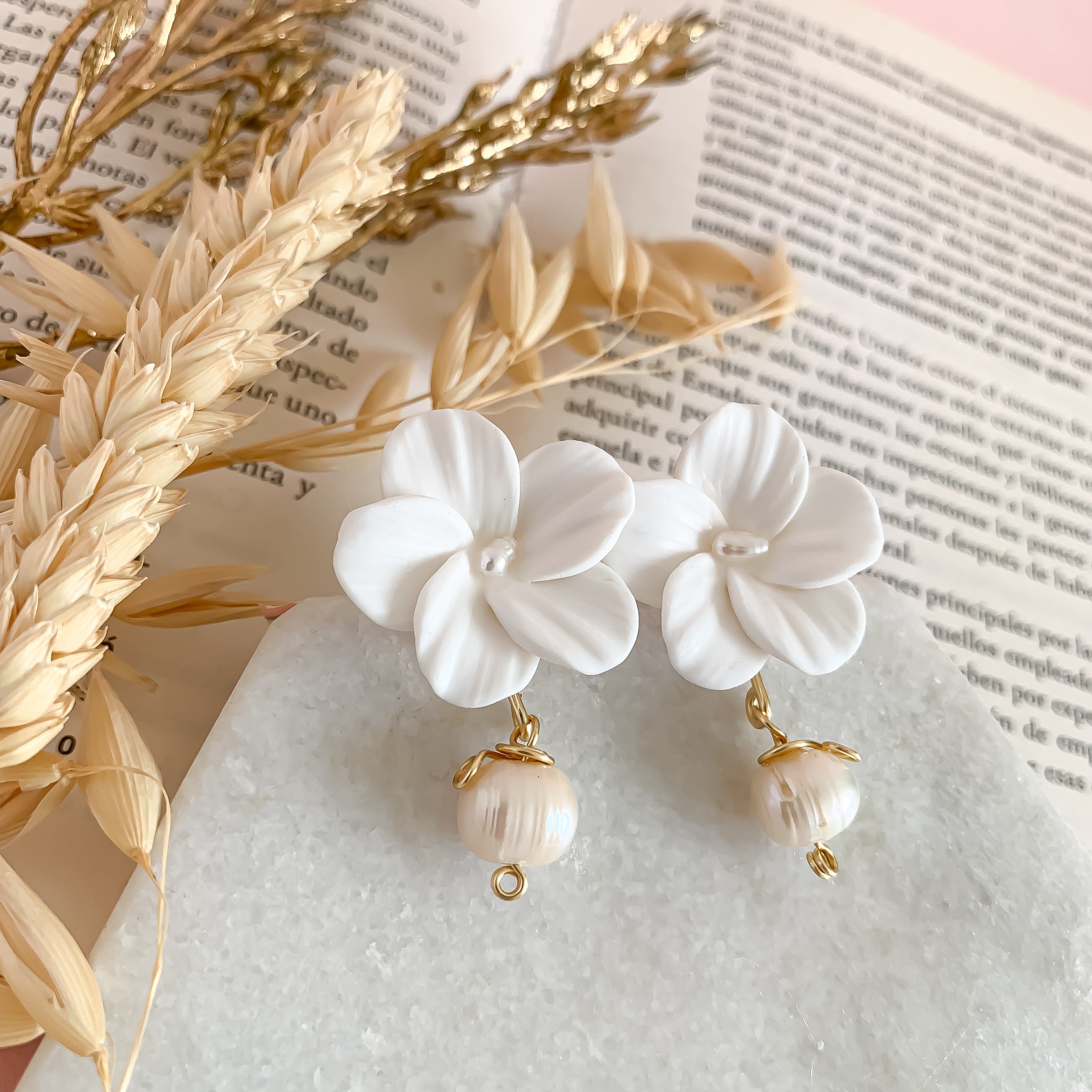 White floral earrings with pearl drops on a textured surface with dried plants and text.