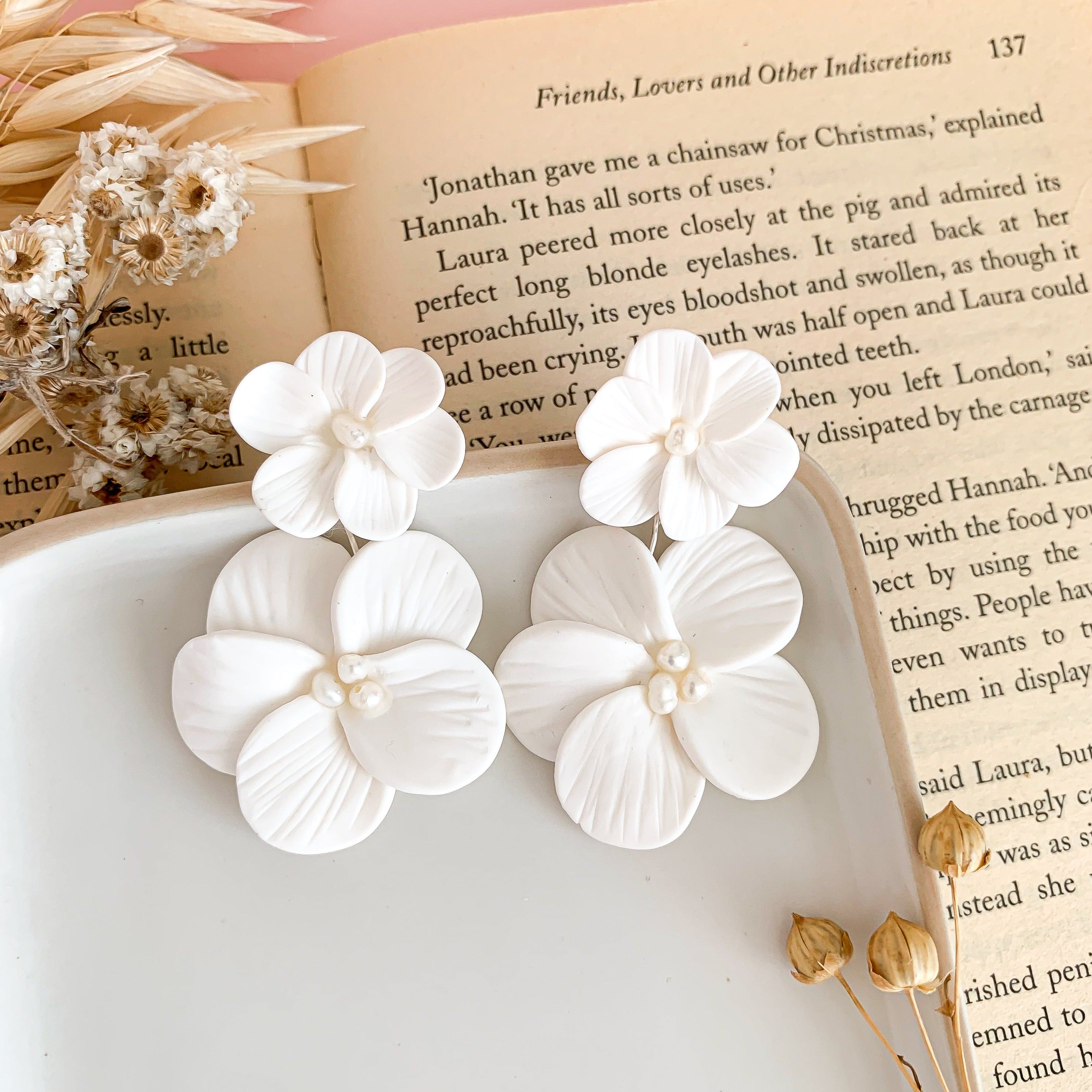 A pair of off-white double flower statement earrings with pearls, displayed on a surface next to an open book and some dried flowers.